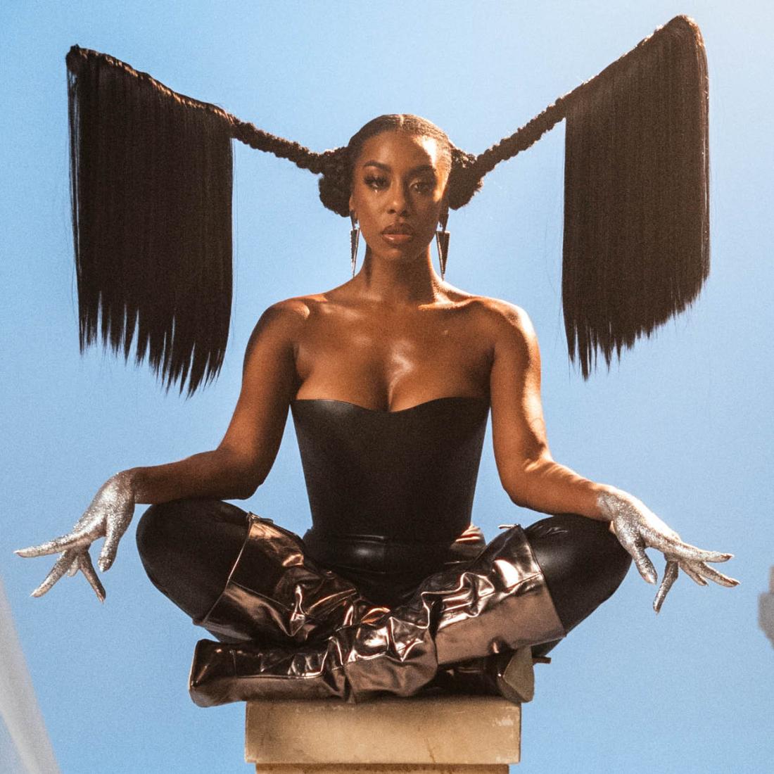 K.ZIA Zit op een platform met een blauw achtergrond. Haar staat in 2 staande vlechten die op het einde los hangen.