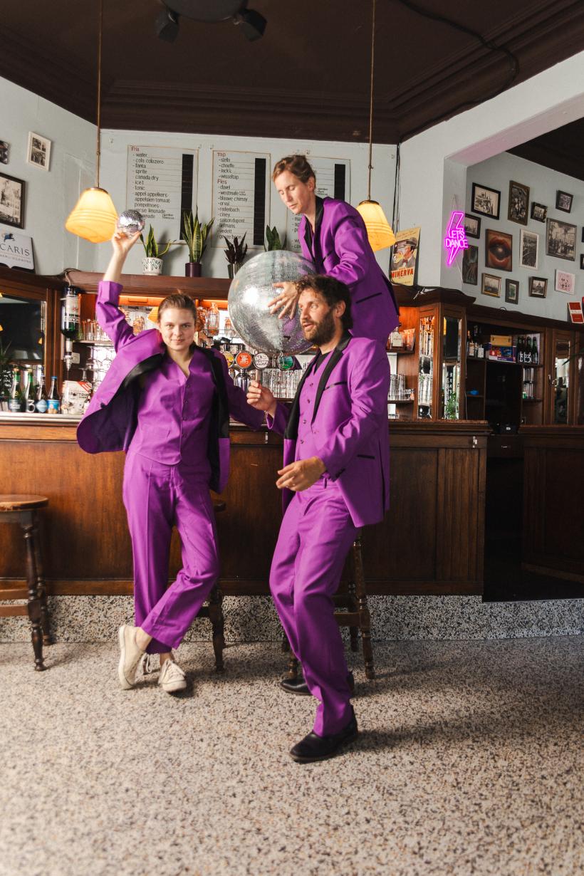 Drie personen (Suzanne Grotenhuis, Freek Vielen en Ellis Meeusen) in paarse pakken die aan een bar staan met een grote discobal. Ze lijken te feesten.