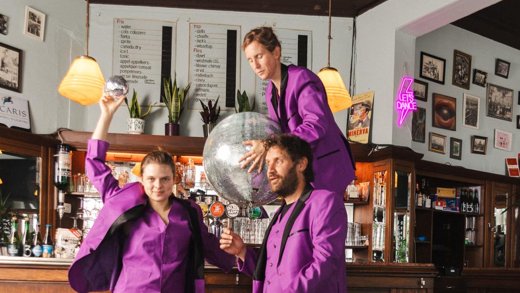 Drie personen (Suzanne Grotenhuis, Freek Vielen en Ellis Meeusen) in paarse pakken die aan een bar staan met een grote discobal. Ze lijken te feesten.